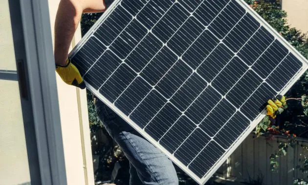 A worker carrying a solar panel for installation on a sunlit rooftop, showcasing clean energy.
