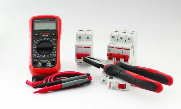Close-up of a digital multimeter with electrical tools and circuit breakers on a white background.