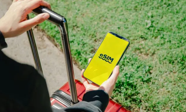 A person holds a smartphone showing an eSIM app while standing with luggage outdoors in Los Angeles.