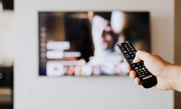 Person using a TV remote control with a blurred television screen in the background.