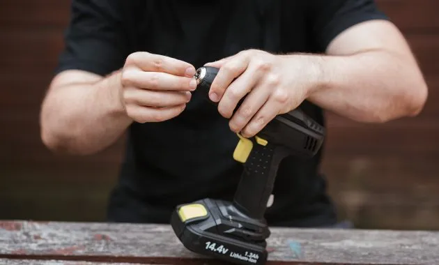 Crop anonymous skilled master in casual outfit preparing electric drill for job on wooden table in soft daylight